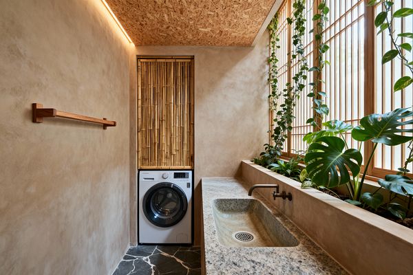 A narrow, light-filled laundry room with warm beige plaster walls and an OSB ceiling with a slim LED strip. A long speckled granite trough sink with a wall-mounted brass tap runs along one side. A white front-loading washer sits below a vertical bamboo screen. Wood-framed slatted windows backlight a built-in planter filled with trailing vines and large monstera leaves. The floor is dark natural stone with white veining, and a simple wood rail mounts on the opposite wall.