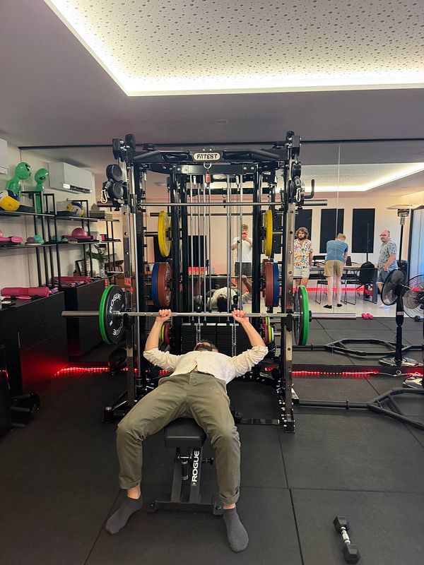 A man with a beard lies on a bench press lifting a barbell in a gym room where three other people interact near a mirror.
