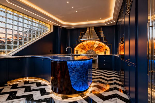 An Art Deco kitchen finished in deep navy cabinetry features a curved island clad in vivid blue stone on a brass plinth, set over a black-and-white chevron marble floor. The cooking wall centers on a stainless range beneath a stepped metallic hood with a dramatic backlit honey onyx sunburst backsplash. Warm cove and toe-kick LED lighting highlight brass trim, while a sloped glass-block window floods the room with daylight, and built-in ovens and a gooseneck faucet complete the luxurious scene.