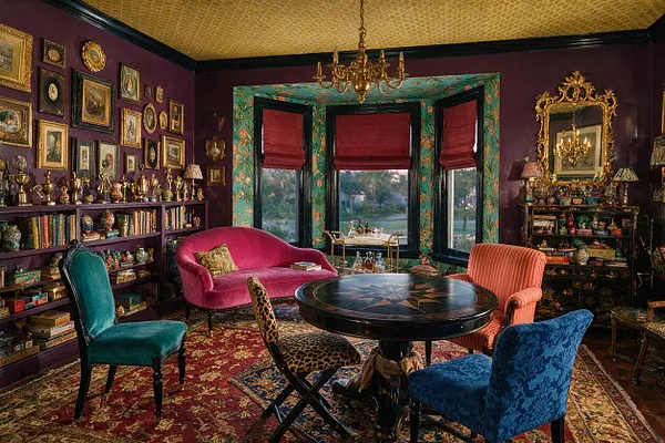 A richly layered game room features deep plum walls, a gold-patterned ceiling, and a round black game table at the center. Around it sit mismatched upholstered chairs in teal, pink, orange, blue, and leopard print, with built-in shelves of books and decorative objects, ornate framed art, gilded mirrors, and a bay window dressed with red roman shades.
