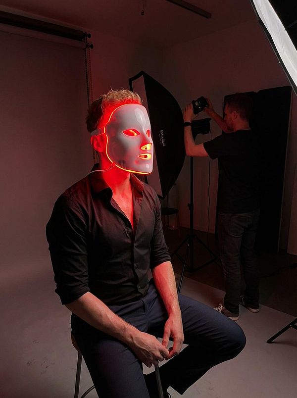 A model wearing a glowing mask sits on a stool in a photography studio.