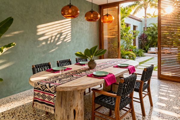 A sunlit tropical dining room features a large oval live-edge wood table on a tree-stump base, surrounded by wooden armchairs with charcoal woven-rope seats. Three amber glass pendant lights hang above a geometric runner with fuchsia napkins and mint plates, while a sage green plaster wall, speckled terrazzo floor, and sliding wood louver doors opening to palm-filled gardens complete the indoor–outdoor setting.