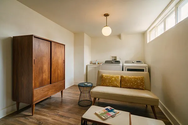 A basement laundry room styled with mid-century modern furnishings, featuring a walnut wardrobe, a tan loveseat with patterned cushions, a small wire side table, and a vintage-style pendant light. A washer and dryer sit against the back wall beneath small high windows, with wood-look flooring and soft neutral walls creating a warm, functional space.