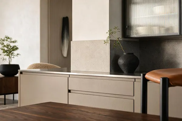 A modern kitchen detail with beige flat-panel cabinets, a dark countertop, and a light stone backsplash beneath ribbed glass upper cabinets. Black ceramic vases, warm wood furniture, and a tan leather stool add contrast to the calm neutral palette.