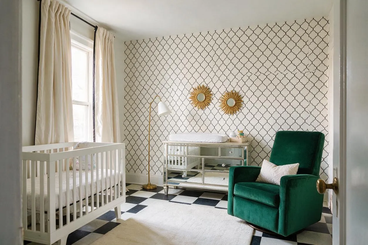 A stylish nursery with a white crib near tall windows draped in cream curtains, a mirrored changing dresser centered on a patterned accent wall, and a deep emerald velvet armchair with a pillow. Gold sunburst mirrors and a brass floor lamp add glamour, while black-and-white checkered flooring and a soft rug ground the space.