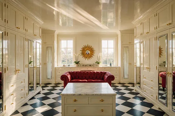 A luxurious walk-in closet with cream built-in wardrobes lining both sides, mirrored panels, and polished brass hardware. A deep red velvet tufted sofa sits at the center behind a marble-topped island, while black-and-white checkered flooring, tall windows, and a gold sunburst mirror complete the Hollywood Regency look.