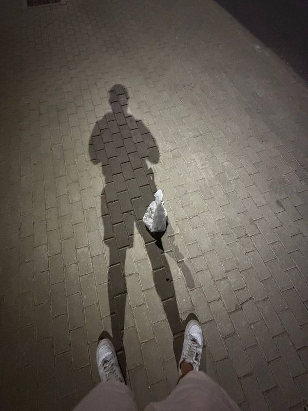Shadow of a person holding a small wrapped item on a brick pavement illuminated by a strong light source.