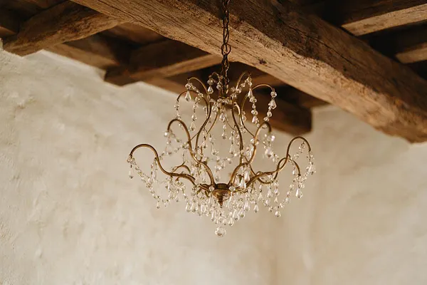 A vintage-inspired chandelier with a dark brass frame and strands of clear crystal drops hangs from a chain beneath thick exposed wooden ceiling beams. The backdrop is a softly textured cream plaster wall, creating a warm French Country contrast between rustic structure and elegant detailing.