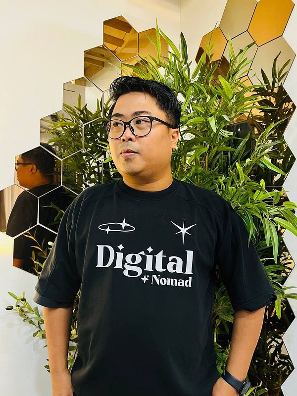 A person wearing a black t-shirt with 'Digital Nomad' printed on it stands in front of a decorative plant backdrop.