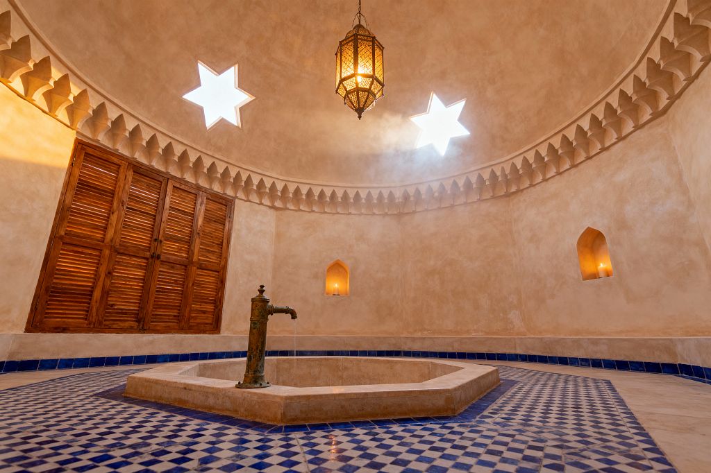 A Moroccan hammam-style bathroom features a domed, sand-colored plaster ceiling punctuated by two star-shaped skylights and a hanging pierced brass lantern. At the center, a sunken stone basin with a patinated brass spout sits on blue-and-white zellige mosaic tiles, while warm tadelakt walls with a sculpted cornice encircle arched candle niches and louvered wooden doors, filling the steamy space with soft, amber light.