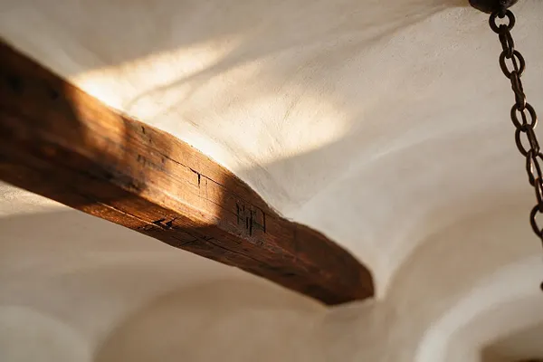 A close-up view of a Mediterranean interior ceiling showing a heavy exposed wooden beam crossing a white plastered vault. Warm sunlight highlights the textured wood grain, and a dark hanging chain appears at the right edge.