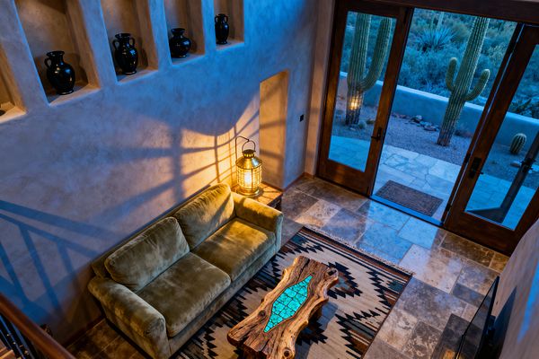 An overhead view of a Southwestern living room shows an olive-green sofa against sand-colored plaster walls with recessed niches holding black pottery. A brass lantern on a side ledge casts dramatic shadows. In front, a live-edge wood coffee table with turquoise stone inlay sits on a geometric wool rug. Travertine tile floors lead to tall wood-framed glass doors opening to a patio and desert landscape with saguaro cacti under cool evening light.