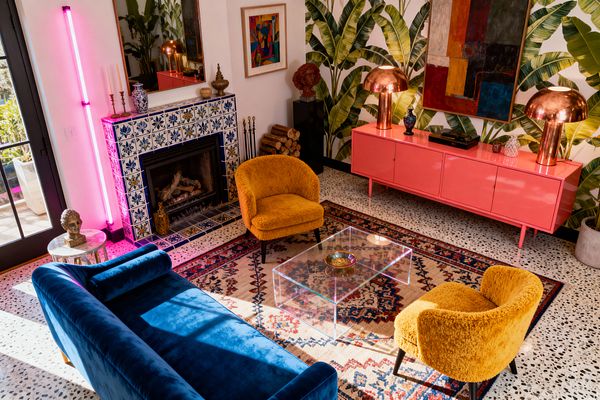 A maximalist living room with a cobalt-blue velvet sofa and two mustard bouclé lounge chairs arranged around a clear acrylic coffee table on a patterned rug over black-and-white terrazzo floors. A blue-and-white tiled fireplace stands beside a vertical hot pink neon light, while a glossy coral credenza holds two dome copper lamps beneath banana-leaf wallpaper; abstract art, a mirror, and black-framed French doors add contrast and daylight.