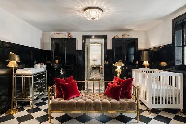 A glamorous nursery with glossy black paneled walls, a black-and-white checkerboard floor, and brass-accented furniture. A tufted velvet daybed with red pillows sits in the center, a white crib stands to the right, and a mirrored changing table and crystal ceiling light add elegance.