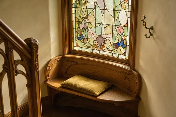 A small Art Nouveau stair landing features a built-in curved wooden bench beneath a stained-glass window. The window shows pastel floral motifs in leaded glass, while the bench has carved organic lines and a mustard cushion, with a decorative wooden railing at left and a simple wall hook on the right.