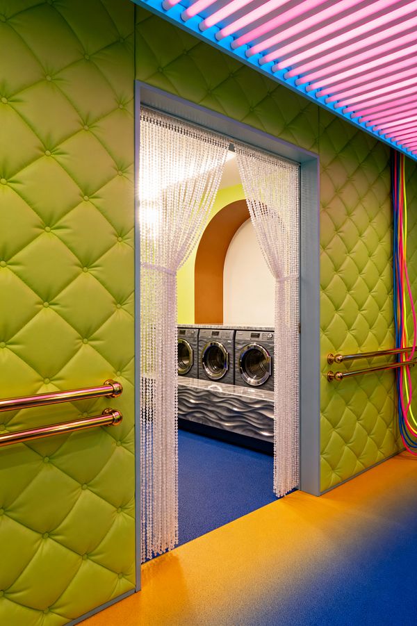 A vivid laundry room framed by lime green, diamond‑tufted padded walls with polished brass handrails and a pale blue doorway. A white acrylic beaded curtain parts to reveal three front‑loading stainless steel washers set in a rippled silver base, backed by a soft green wall and an ochre arched niche. The floor shifts from golden orange to electric blue, while linear pink‑and‑blue neon tubes light the ceiling; multicolored cables hang along the right wall.
