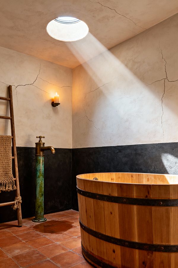 A Wabi-Sabi bathroom features a cedar soaking tub bound with dark metal bands, lit by a round skylight that sends a beam across hand-troweled lime-plaster walls with visible hairline cracks and a two-tone finish, warm off-white above and charcoal below. A tall patinated brass column faucet, a lit candle sconce, terracotta floor tiles, and a rustic wooden ladder draped with a chunky woven jute towel complete the restrained, timeworn setting.
