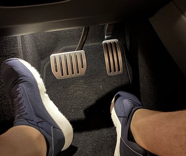 Close-up of a Tesla Model 3 aluminum pedal installed next to the original black plastic pedal on the car's footwell floor.