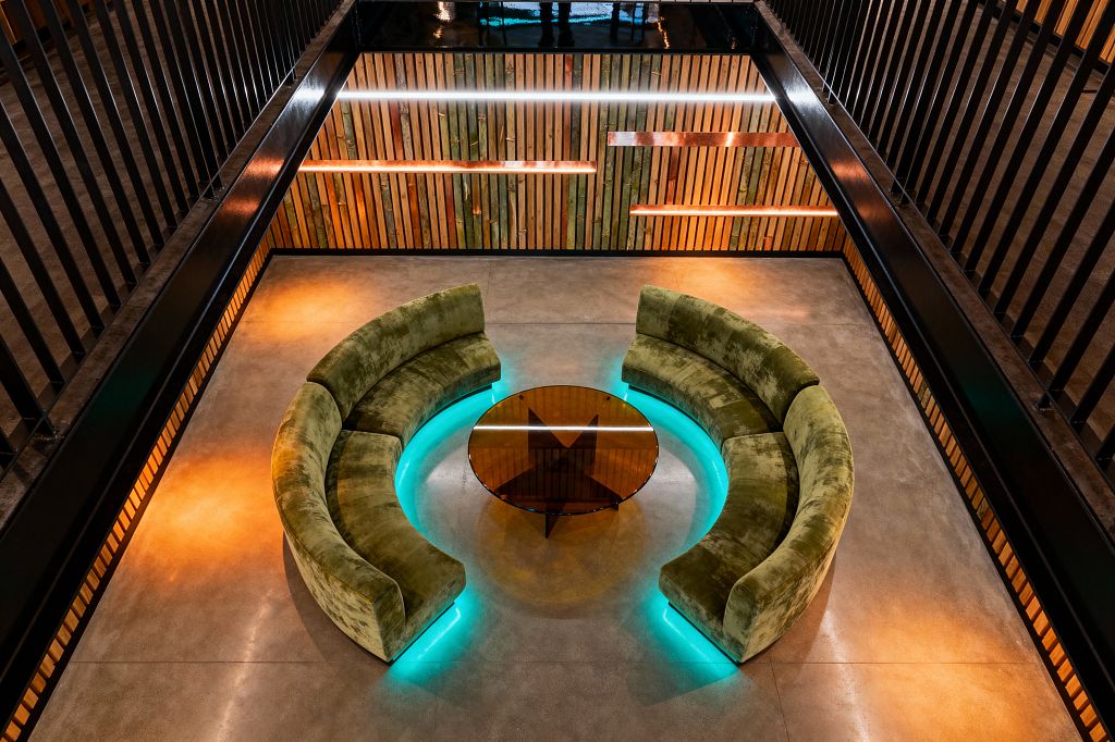 Top-down view of a futuristic living room featuring two semicircular moss-green velvet sofas forming a conversation pit around a round amber-tinted glass table. Cyan LED lighting runs beneath the inner edges of the sofas, illuminating a polished concrete floor, while a vertical slatted wood feature wall with integrated linear light bars and black steel railings defines the surrounding architecture.