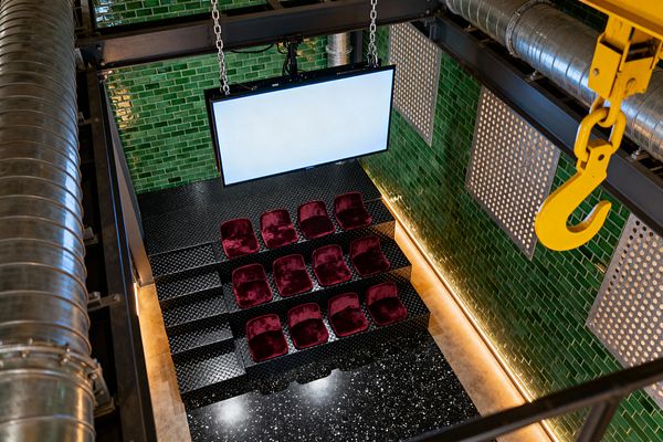Overhead view of an industrial-style media room with emerald green glazed brick walls and a large flat screen suspended by chains. Three rows of burgundy velvet bucket seats sit on black diamond-plate steel risers above a black terrazzo floor, surrounded by exposed steel beams, galvanized ductwork, perforated metal panels, and a bright yellow crane hook, with warm linear LED lighting washing the base of the wall to create a moody, theater-like atmosphere.