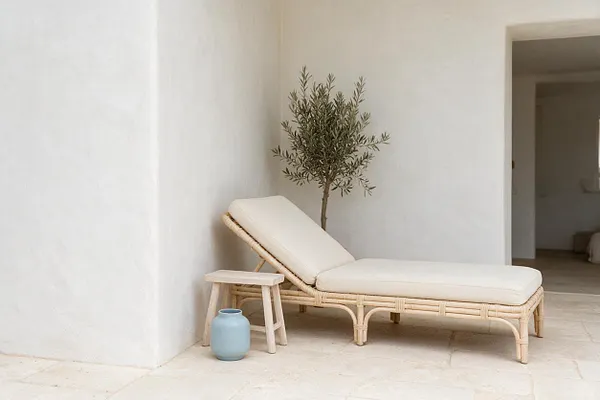A minimalist coastal exterior patio with white stucco walls and pale stone flooring. A woven rattan chaise lounge with cream cushions sits beside a small wooden stool and a light blue ceramic vase, with a slender olive tree adding a natural accent.