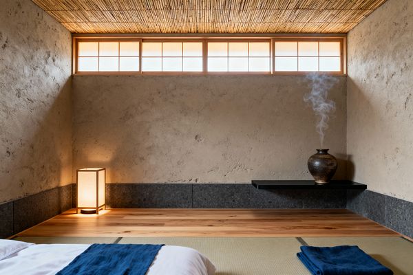 A tranquil Japanese-style bedroom features tatami mats and a low futon with a deep indigo throw in the foreground. Along the back wall, a raised cedar platform holds a paper lantern, while a floating black shelf displays a ceramic vase releasing incense smoke. Textured beige plaster walls sit above a charcoal stone base, and a warm clerestory shoji window spans the room under a bamboo reed ceiling, bathing the space in soft, diffused light.