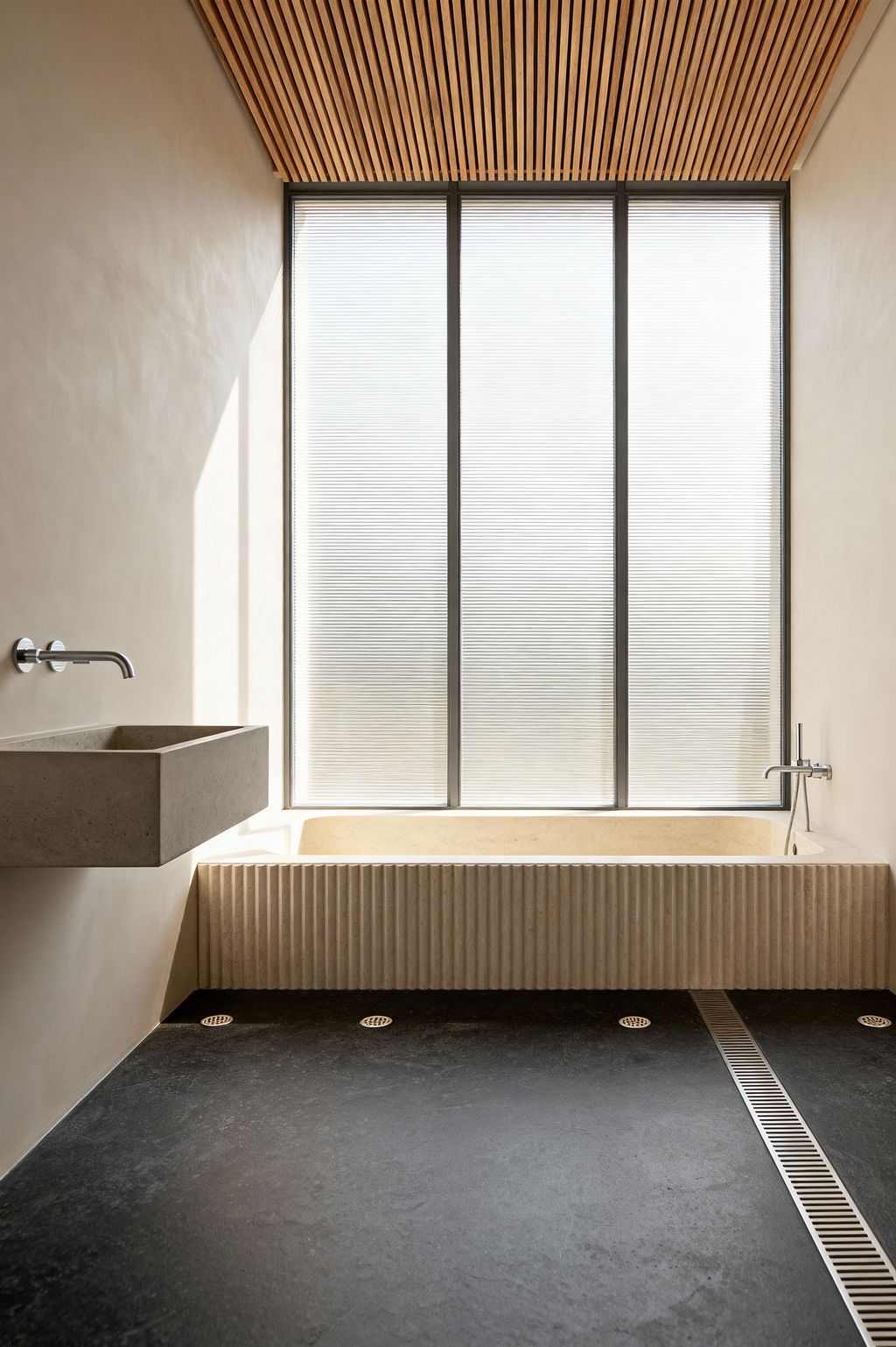 A minimalist bathroom featuring a wall of reeded translucent glass that diffuses daylight. A pale limestone soaking tub with a vertically fluted front sits beneath the window with a wall-mounted chrome filler, while a floating rectangular cast-concrete sink and spout occupy the left wall. The ceiling is lined with natural oak slats, and the floor is honed black stone with a stainless linear drain and round grates, framed by smooth warm off-white plaster walls.