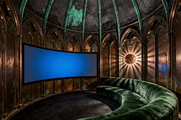 An immersive media room with a ribbed, domed ceiling and walls clad in patinated copper panels detailed with Gothic pointed arches. A large curved projection screen faces a semicircular deep green velvet sofa on dark charcoal carpet. A round bronze sconce emits sunburst rays, washing warm light over the aged metal. Copper, verdigris, black, and forest green set a moody, dramatic atmosphere for cinematic viewing.