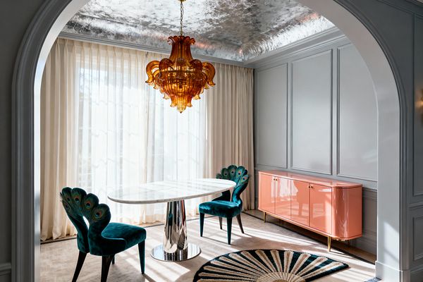 A dining room framed by an arched opening features a white oval marble table on a polished chrome pedestal, flanked by two teal velvet chairs with peacock‑feather backs. An amber glass chandelier hangs beneath a silver‑leaf ceiling, with pale gray paneled walls, floor‑to‑ceiling ivory sheers, a coral lacquer credenza with brass legs, light wood flooring, and a round black‑and‑cream sunburst rug completing the glamorous space.