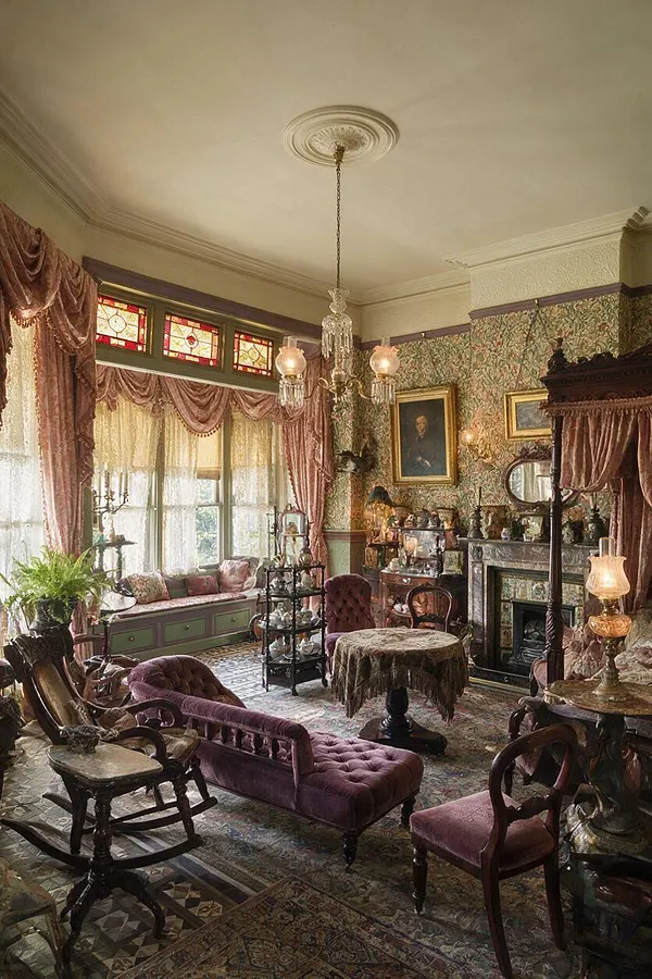 A richly decorated Victorian-style room with patterned wallpaper, stained glass transom windows, heavy draped curtains, and a central crystal chandelier. Plush plum velvet seating, carved wood furniture, a patterned area rug, and an ornate fireplace create a formal, antique-filled atmosphere.