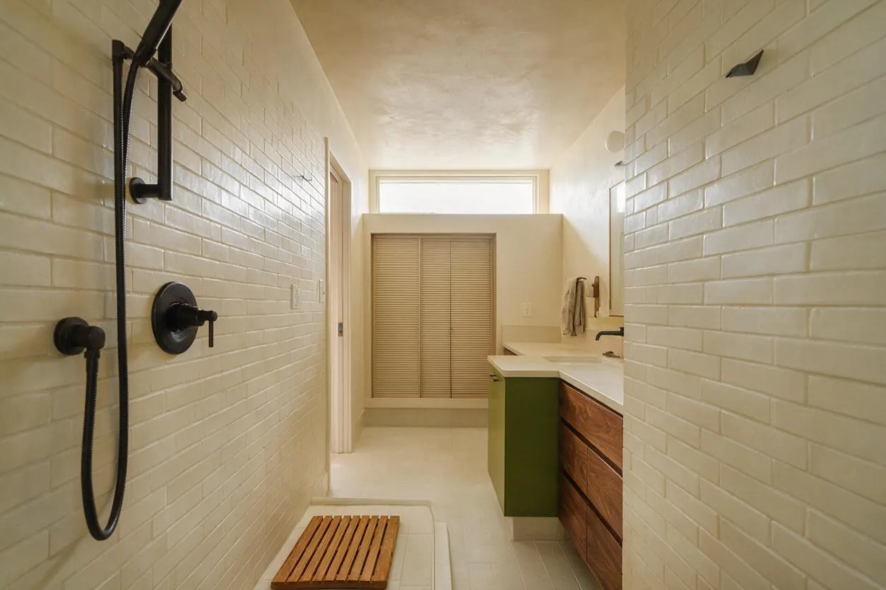 A narrow master bathroom with white subway tile walls, matte black shower fixtures, and a low shower floor with a wooden slatted mat. At the far end, a vanity with walnut drawers, an olive green side panel, and a light countertop sits beneath a mirror, with a small window and louvered closet door beyond.