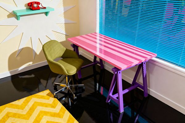 A Pop Art home office features a glossy hot pink desk on purple trestle legs beside a large window with bright blue Venetian blinds casting striped shadows across the surface. An olive green upholstered swivel chair with a chrome base sits on a sunny yellow chevron rug over a high-gloss black floor; pale yellow walls show a white starburst graphic and a mint floating shelf topped with a red rotary telephone, creating a bold, comic-book-inspired palette.