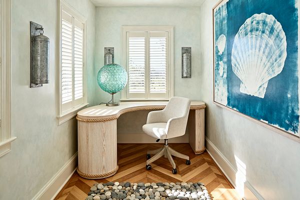A compact coastal home office centers on a curved, whitewashed wood desk wrapped with thick rope beneath windows with white plantation shutters, paired with a beige upholstered swivel chair on warm herringbone oak flooring and a pebble-stone rug. Marine details—the bubbled aqua glass table lamp, galvanized metal wall sconces, and a large cyan-blue shell artwork—contrast soft seafoam walls and abundant daylight for an airy, beachy feel.