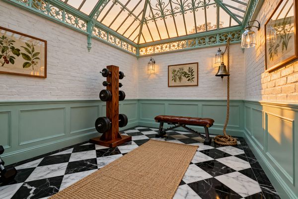 Traditional home gym set in a conservatory with a pitched glass roof and mint-green wrought-iron detailing, surrounding white-painted brick and matching mint wainscoting above a black-and-white marble checkerboard floor layered with a natural sisal runner. A brown leather weight bench and wood dumbbell stand with cast-iron plates sit centrally as botanical prints, clear-glass lantern sconces, and a wall-mounted brass bell with a thick rope glow in warm evening light.