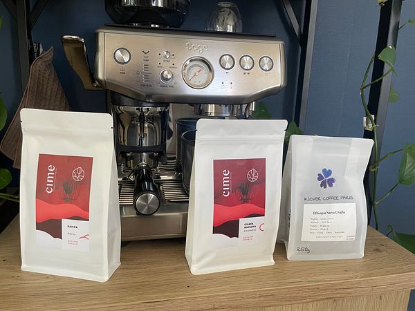The image features three bags of specialty coffee in front of an espresso machine.