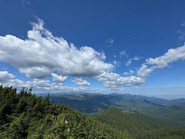 A scenic mountain landscape with a person hiking along a rocky trail surrounded by some green shrubbery and small trees under a partly cloudy blue sky.