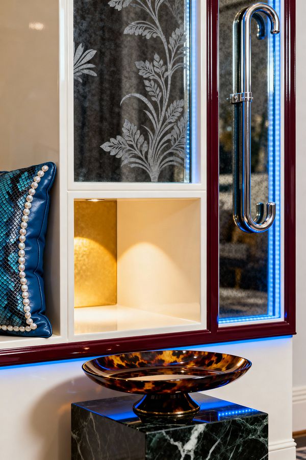 Close-up of a Hollywood Regency mudroom built-in with glossy white cubbies framed in burgundy lacquer. One niche has a gold-leaf back and warm downlight; adjacent panels feature antiqued mirror with etched botanical motifs and blue LED perimeter lighting. A teal faux snakeskin bench cushion with pearl trim sits at left. Below, a dark green marble pedestal holds a tortoiseshell bowl, with a polished chrome handle at right.