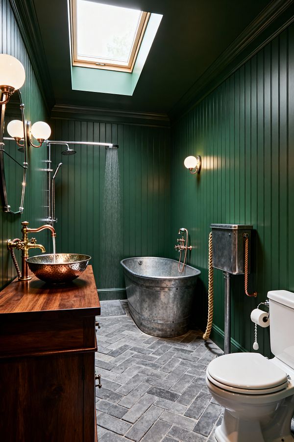 A narrow bathroom with dark green beadboard walls and ceiling is lit by a skylight. A walnut vanity holds a hammered copper vessel sink with exposed brass plumbing. At the back, a galvanized steel soaking tub sits under a wall-mounted rainfall shower, while warm opal globe sconces flank the space. On the right, a white toilet pairs with a high-level metal cistern and rope pull. The floor is finished in charcoal gray herringbone brick tile.