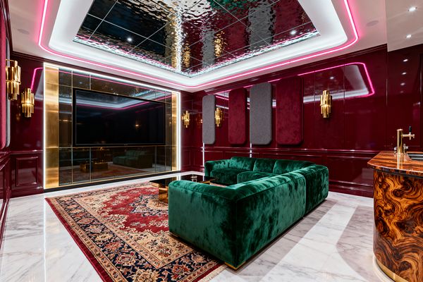 A glamorous media room features glossy burgundy paneled walls with brass sconces and a wall-mounted TV framed by brass-trimmed mirrored panels. An emerald green velvet sectional sits on a red Persian-style rug over white marble flooring, facing a black glass coffee table; burgundy and charcoal acoustic panels line the walls, a rippled mirrored ceiling with pink and white cove LEDs adds glow, and a curved burl-wood bar with a brass faucet occupies the right corner.