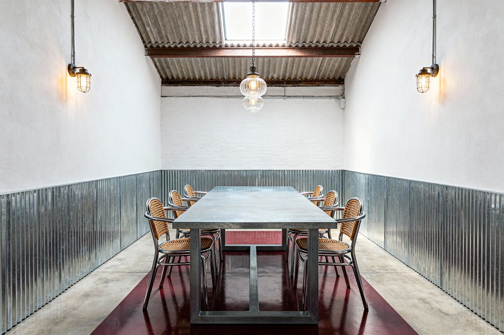A long rectangular metal dining table sits centered on a deep oxblood floor inset, surrounded by eight aluminum-framed chairs with woven rattan seats and backs. White plaster walls capped with corrugated steel wainscot lead to a white brick end wall, under a corrugated roof with exposed beams and a skylight; a ribbed-glass pendant and two cage bulkhead sconces provide warm light.