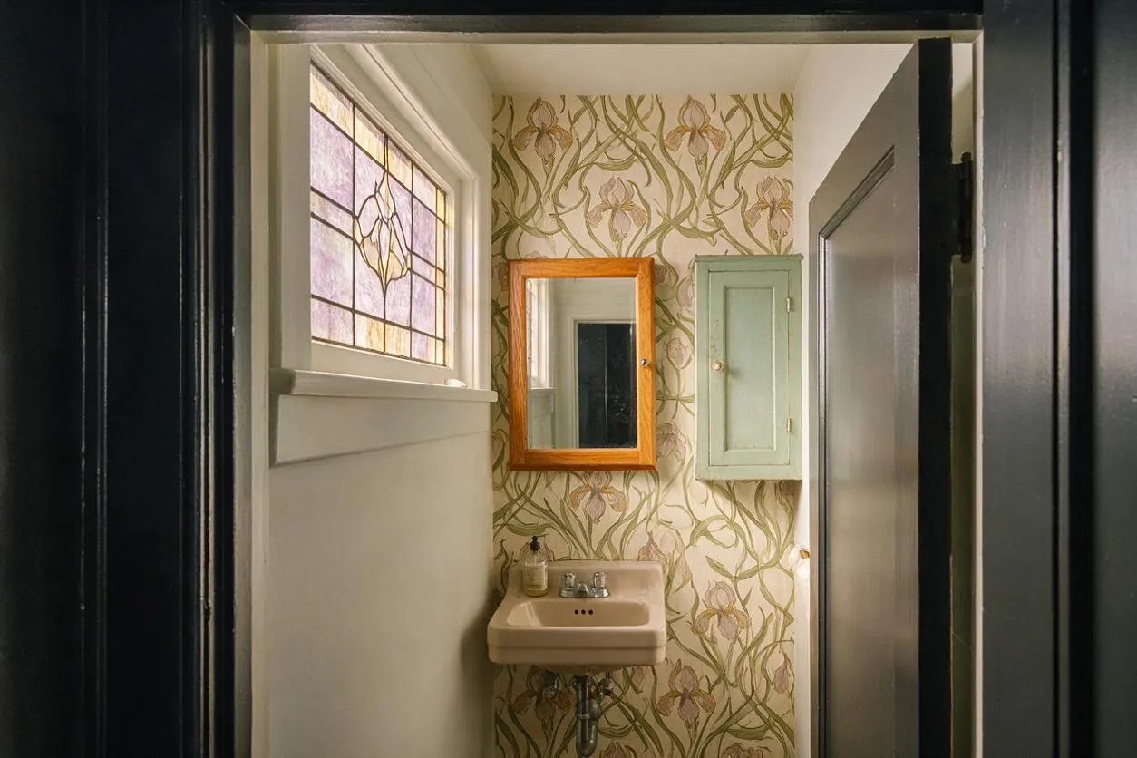A narrow powder room with Art Nouveau floral wallpaper, a small beige wall-mounted sink, and a wood-framed mirror above it. A stained-glass window on the left adds soft colored light, while a pale mint medicine cabinet and dark doorway trim complete the vintage look.