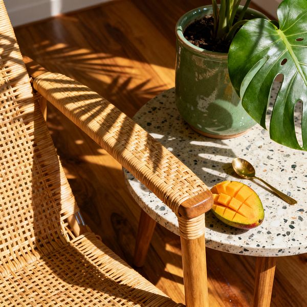 Close-up of a tropical patio corner with a woven rattan chair beside a round terrazzo side table on warm wood flooring. A green glazed pot with a monstera plant, a halved scored mango, and a small gold-toned spoon sit in dappled sunlight, with palm-leaf shadows across the chair and tabletop, emphasizing natural textures and a relaxed, sun-washed atmosphere.