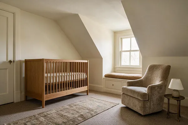 A serene transitional nursery with a natural wood crib, a cushioned window seat beneath a bright window, and a soft taupe armchair beside a small round side table and lamp. The room has sloped ceilings, cream walls, beige carpet, and a muted patterned runner rug.