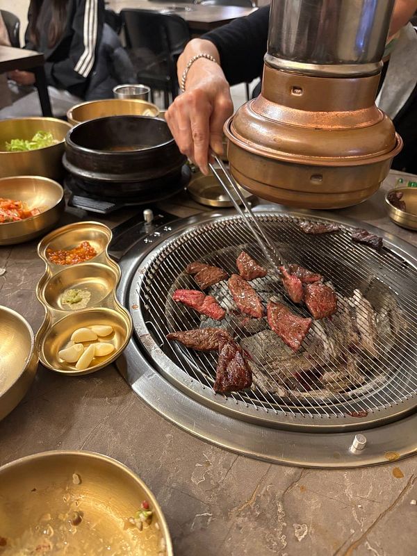 A person grilling pieces of beef on a Korean BBQ charcoal grill surrounded by various traditional side dishes.