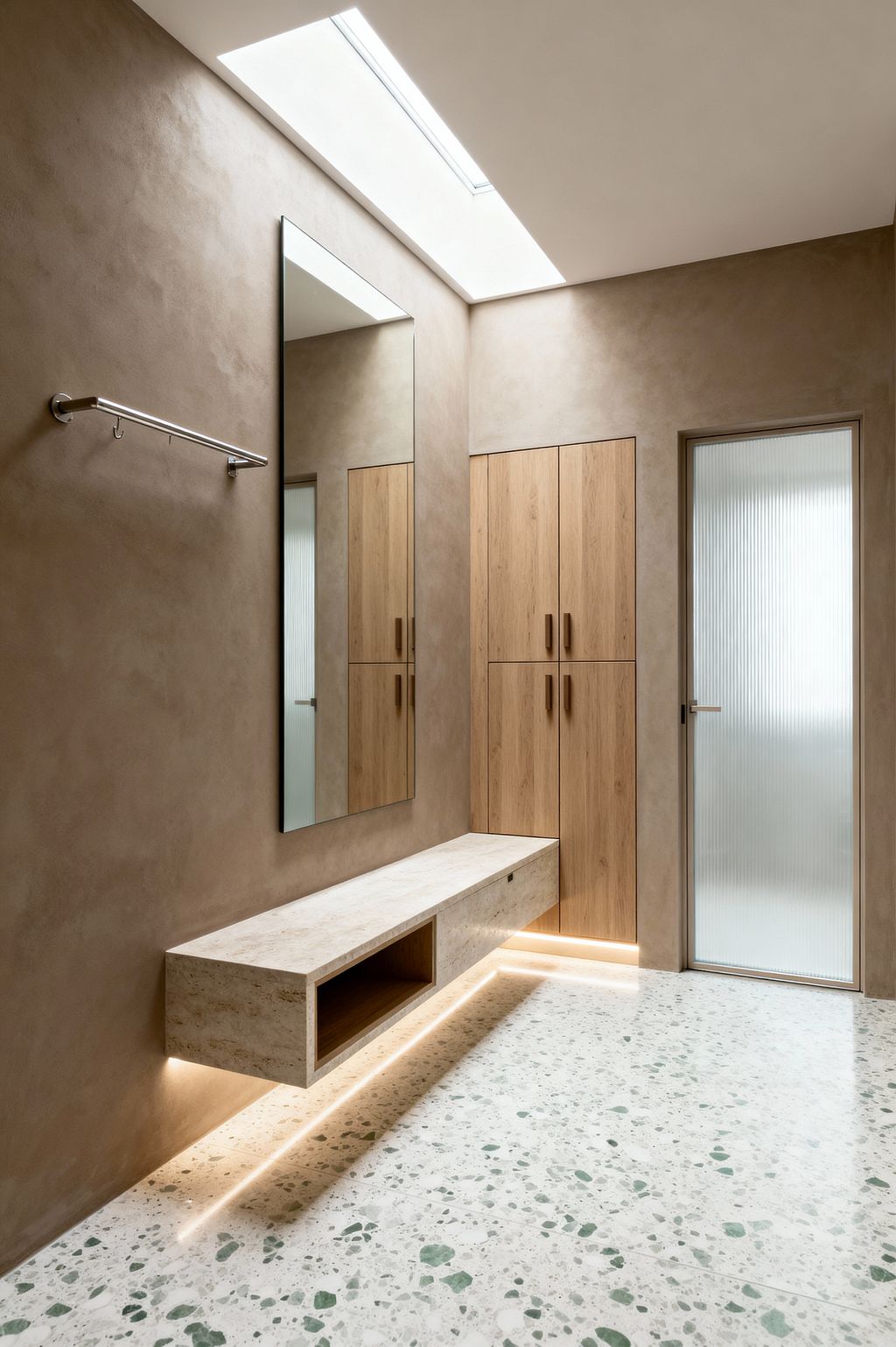 A minimalist mudroom with taupe plaster walls and a white terrazzo floor with green flecks. A floating travertine bench with an open cubby glows with under-mounted LED lighting, facing a tall frameless mirror and a slim stainless steel rail with hooks. Built-in oak cabinets line one wall, and a ribbed glass door and long skylight flood the space with soft, diffused light.