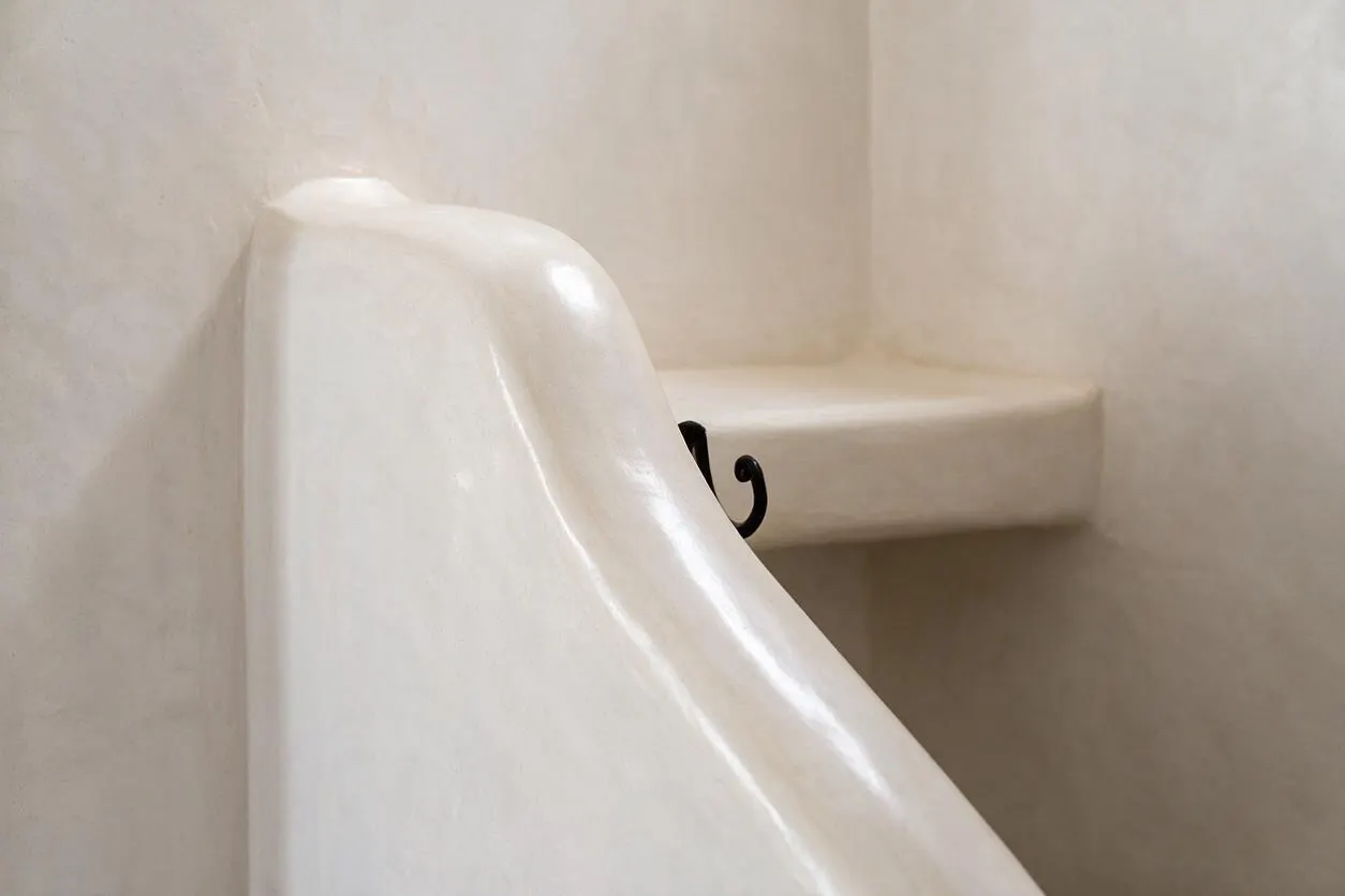 A close-up of a Moroccan-style staircase finished in smooth glossy white plaster, with a curved built-in handrail or stair edge and a soft beige plaster wall and landing behind it. A small black wrought-iron detail peeks from the side, adding contrast to the minimalist organic form.