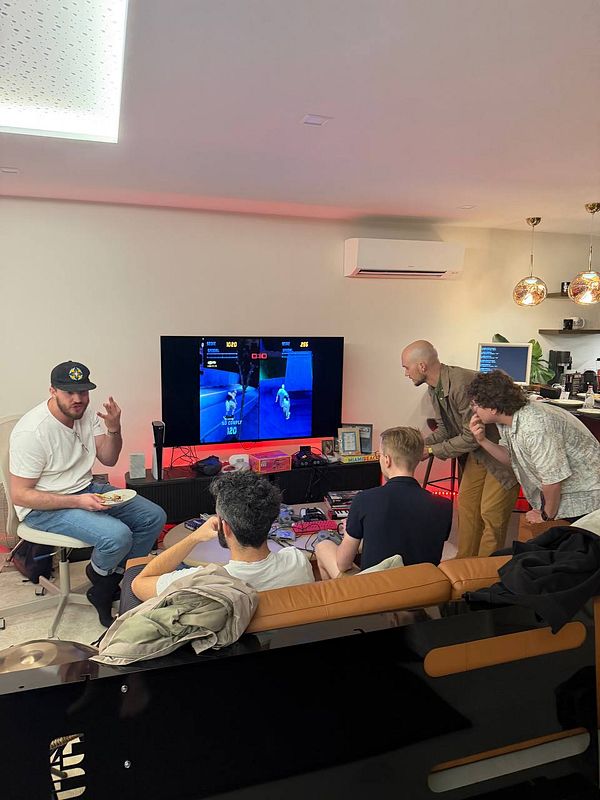 Five men in a casual coworking space are gathered around a TV playing a video game, with some sitting and some standing, engaging in the game and conversation.