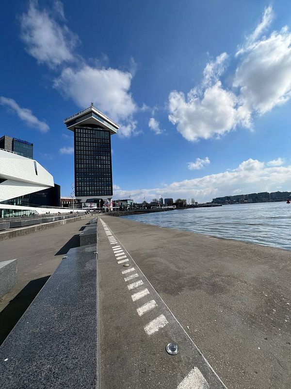 A modern building stands beside a calm waterfront under a clear blue sky with scattered clouds.