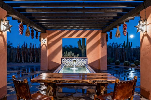 An outdoor patio under dark wooden pergola beams features terracotta stucco walls and a long, blue-and-white Talavera-tiled fountain centered beneath an open window that frames desert cacti and a full moon. Dried red chile ristras hang near star-shaped metal-and-glass lanterns, while a live-edge wood table with stitched leather chairs sits on wet flagstone reflecting the warm light.