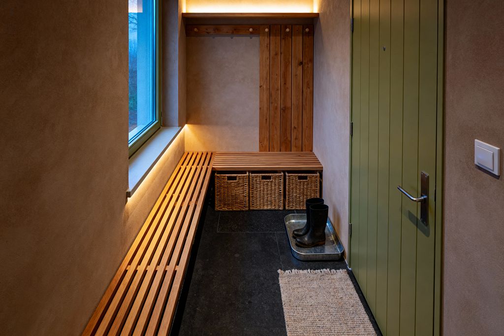 Compact mudroom with warm beige plaster walls and a sage green, vertical‑plank door. A long slatted wood bench runs beneath a window and turns into a corner seat above three woven baskets. Vertical timber boards with simple pegs form a hanging area. Concealed LED strips glow under the windowsill and shelf, lighting dark slate flooring. A jute runner and a galvanized tray with black boots underscore functional Scandinavian simplicity.
